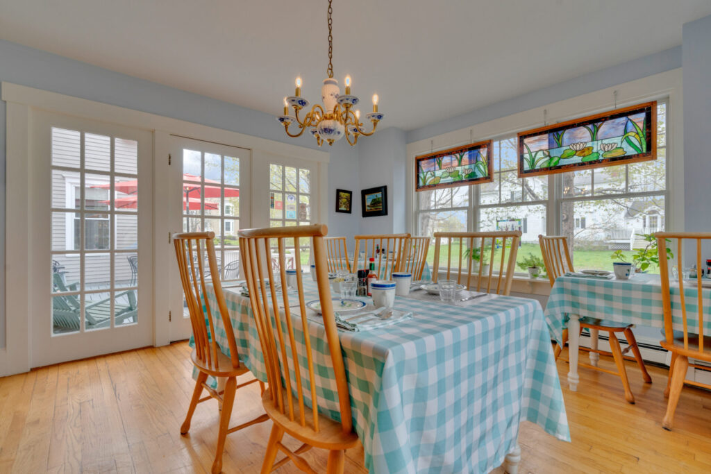 Dining room with wooden chairs, blue checkered tablecloths, chandelier, and stained glass windows facing a garden.