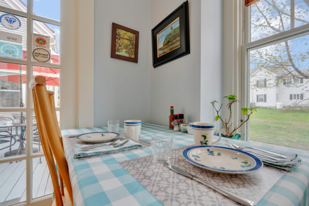 Small dining table set for two by a window, with dishes, mugs, and condiments on a blue and white checkered tablecloth.