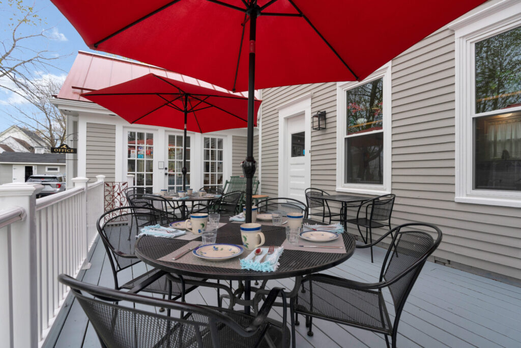 Outdoor patio with black metal tables and chairs, set with dishes, under large red umbrellas beside a gray house.