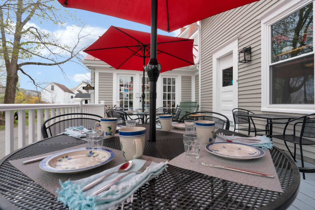 Outdoor table set for four with plates, cups, and utensils under red umbrellas on a patio beside a gray house.