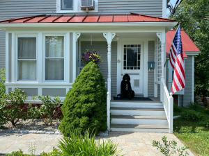 A black dog sits on the porch of a gray house with a red roof and an American flag displayed near the steps.