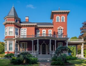 A large red Victorian-style house with a tower and wraparound porch, perfect for autumn in Maine, set against a blue sky.