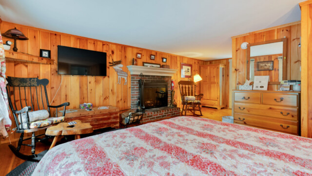 Cozy room with wood paneling, a brick fireplace, TV, rocking chairs, a dresser with a mirror, and a floral bedspread.