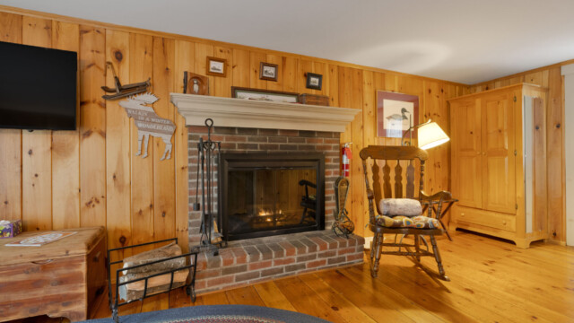 Cozy room with a wooden interior, brick fireplace, rocking chair, and a TV. Rustic decor and ambient lighting.