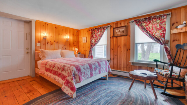 Cozy bedroom with wood-paneled walls, a bed with a red patterned quilt, a rocking chair, and two windows with floral curtains.