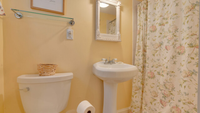 A bathroom with a pedestal sink, toilet, floral shower curtain, white framed mirror, and a small basket on the toilet tank.