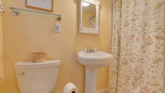 A bathroom with a pedestal sink, toilet, floral shower curtain, white framed mirror, and a small basket on the toilet tank.