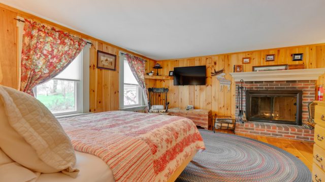 Cozy bedroom with wood-paneled walls, a brick fireplace, floral curtains, a TV, and a bed with a striped quilt.