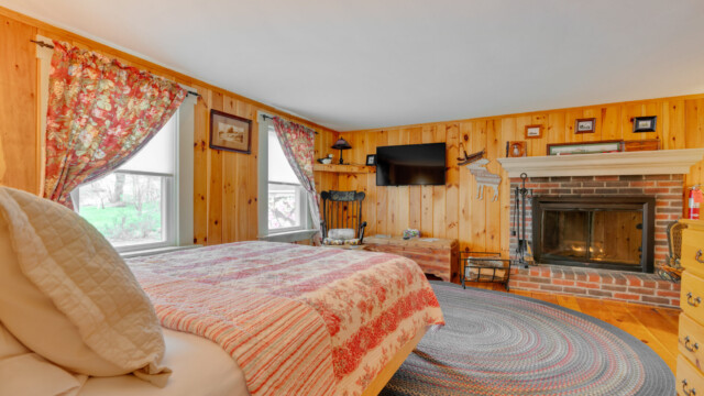 Cozy bedroom with wood-paneled walls, a brick fireplace, floral curtains, a TV, and a bed with a striped quilt.