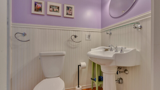 A small bathroom with a purple wall, white toilet and sink, round mirror, three small pictures, and wooden floor.