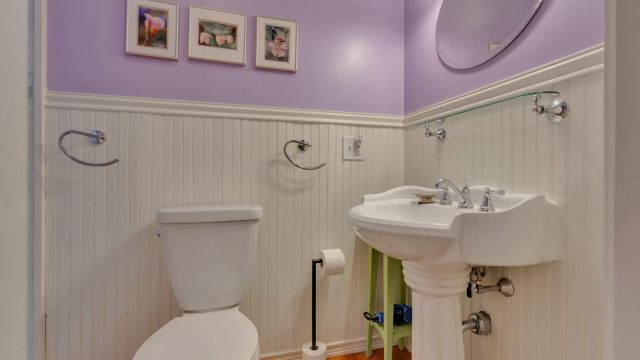 A small bathroom with a purple wall, white toilet and sink, round mirror, three small pictures, and wooden floor.