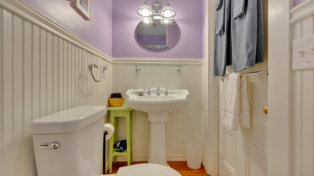Show thumbnail preview A small bathroom with lilac walls, a white pedestal sink, a toilet, towels on a rack, and a round mirror above the sink.