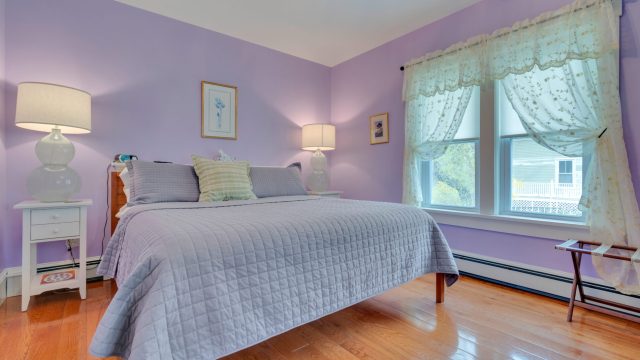 A bedroom with a purple bedspread, bedside tables with lamps, and lace curtains on the window, featuring purple walls.