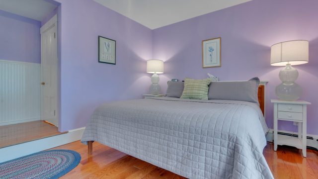 A cozy bedroom with lavender walls features a neatly made bed, two bedside lamps, a rug, and framed art on the walls.