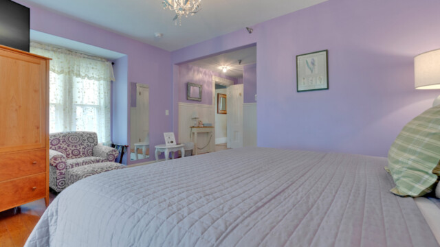A bedroom with purple walls, a bed, armchair, lamp, chandelier, and dresser with a mirror by the window.