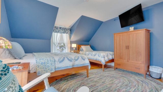 Room with two single beds, blue walls, a wooden armoire, and a TV mounted above. A braided rug covers the wooden floor.