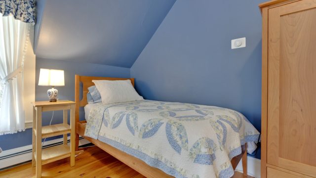 A cozy bedroom with a single bed, blue walls, a wooden nightstand, and a lamp next to a window with floral curtains.