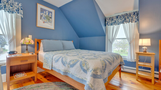 A cozy bedroom with blue walls, wooden bed, and two windows with white curtains. A quilted bedspread and a lamp are present.