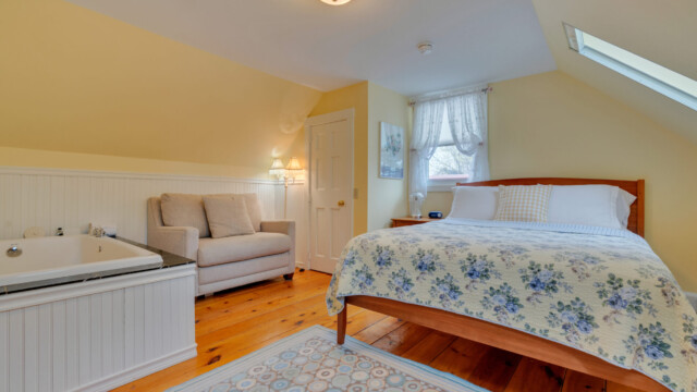Cozy bedroom with a bed, armchair, and a built-in tub. Sloped ceiling with skylight and a soft yellow wall.