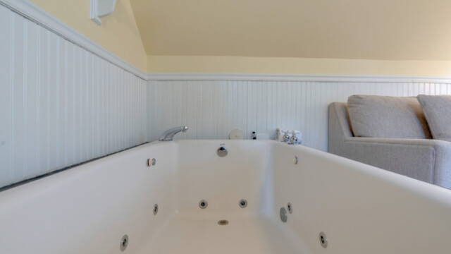 A large white jacuzzi tub with jets in a bathroom, next to a gray couch against light yellow walls and white paneling.