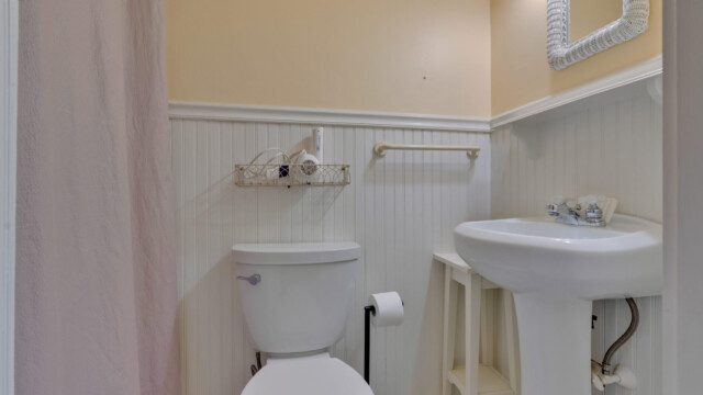 Bathroom with a white toilet, pedestal sink, beige walls, and beadboard paneling. A small shelf holds toiletries above the toilet.